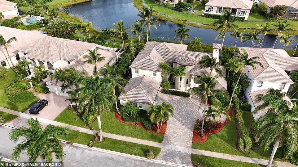 The magnificent home sits amid towering palm trees in a gated community. A balcony rests above an entrance archway, which is beside a rounded turret that appears at least three levels high