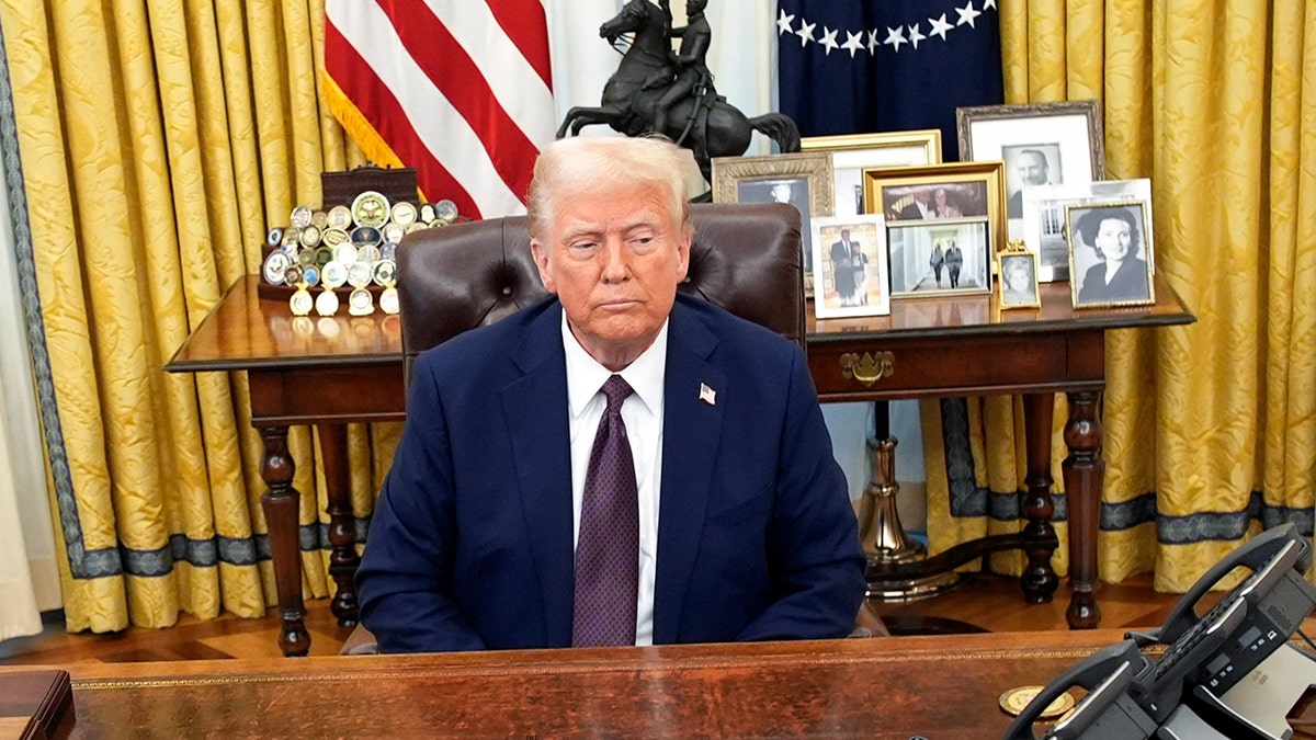 Predsjednik Trump sjedi za odlučnim stolom s obiteljskim fotografijama iza njega.