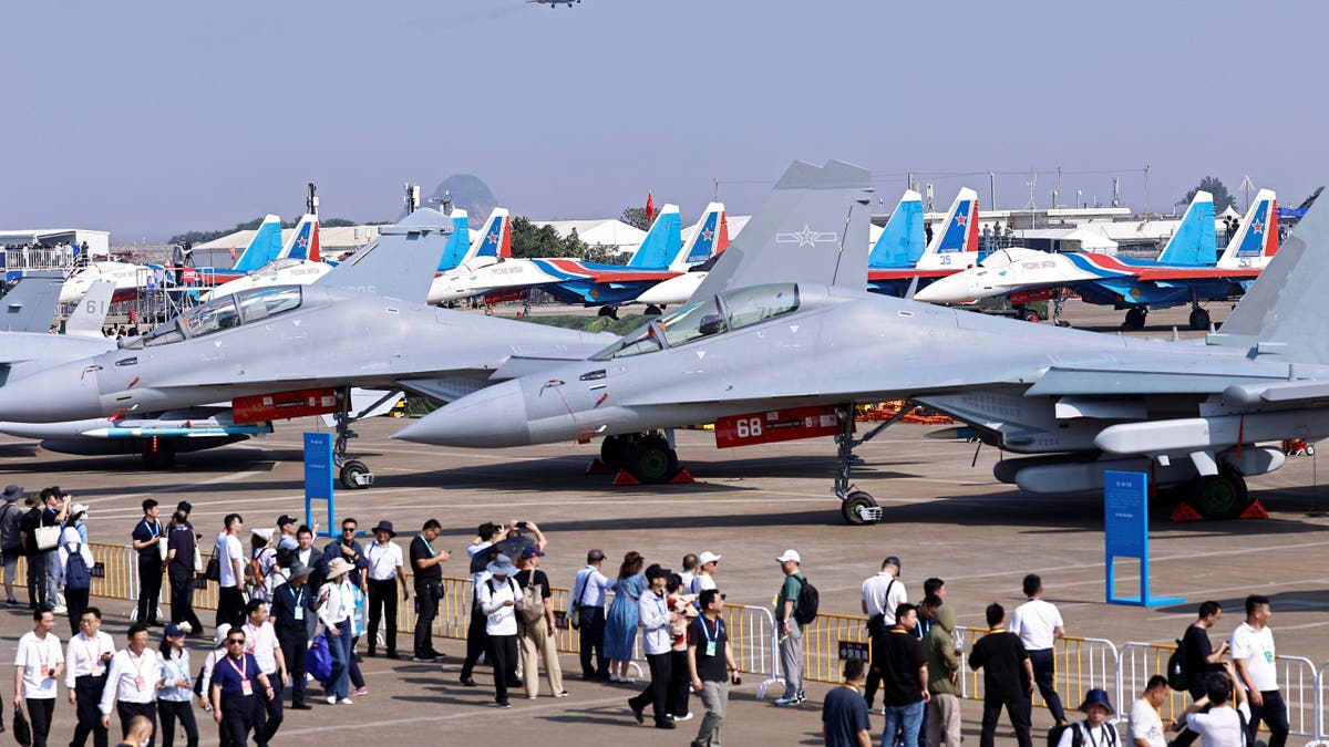 Gužve posjetitelja prolaze pokraj nekoliko borbenih mlaza J-15 kineske mornarice PLA na statičkom prikazu u Airshow China 2024, s drugim zrakoplovima i mlazom koji lete iznad glave.