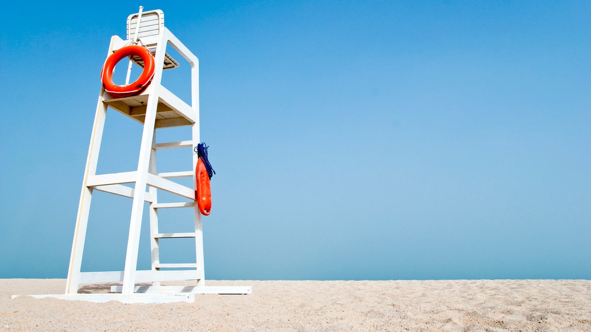 Prazna bijela stolica za spasilac na praznoj plaži s narančastim životnim plutačima visi sa strane. Stolica stoji protiv bistrih plavog neba u pijesku.
