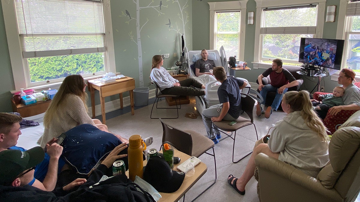 Markus Schouten, seated in a hospital bed, is surrounded by a group of friends and family in a sunlit room.