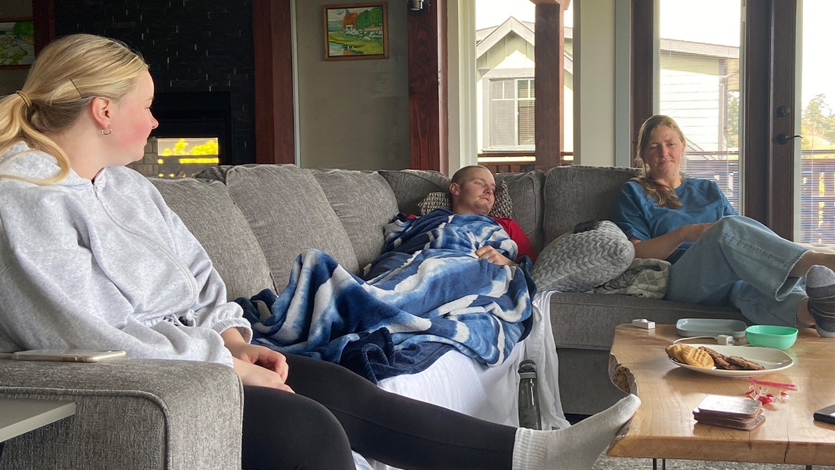 Markus Schouten lies on a gray sectional couch under a blue-and-white blanket near two women