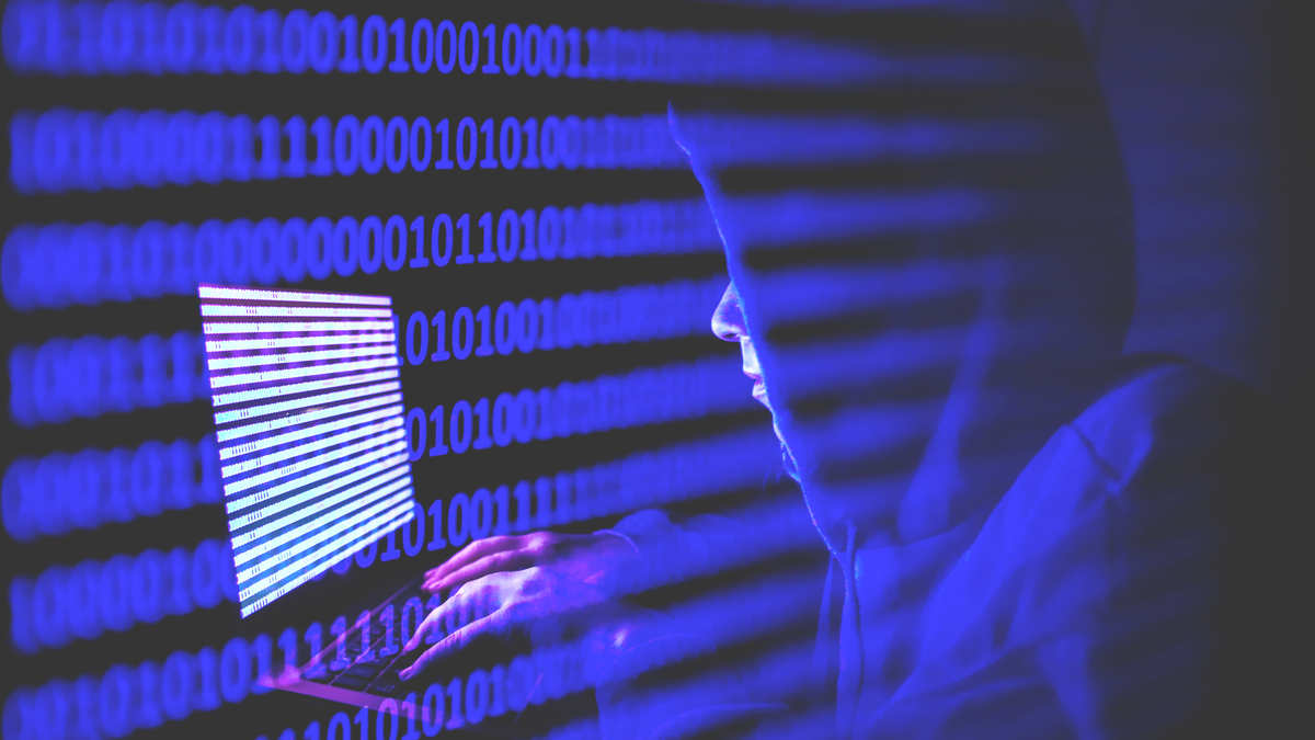 Osoba odjevena kao internetski haker radi na prijenosnom računalu s binarnim kodom na ekranu.