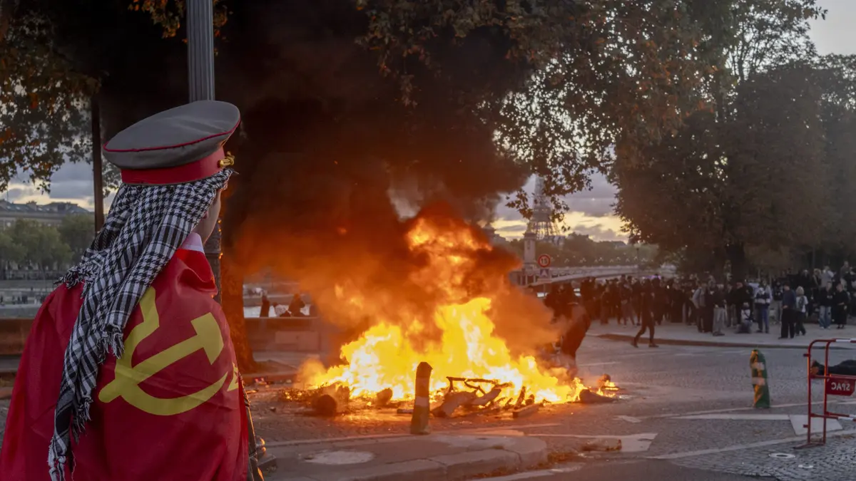 A protester wearing a hammer-and-sickle flag watches a fire burn at a demonstration in Paris, France, on Saturday, Oct. 17, 2026. Rioters attacked French public buildings and looted stores in cities including Paris as protests against the high cost of living drew more than half a million people, underscoring the nation's challenges ahead of presidential elections next year.